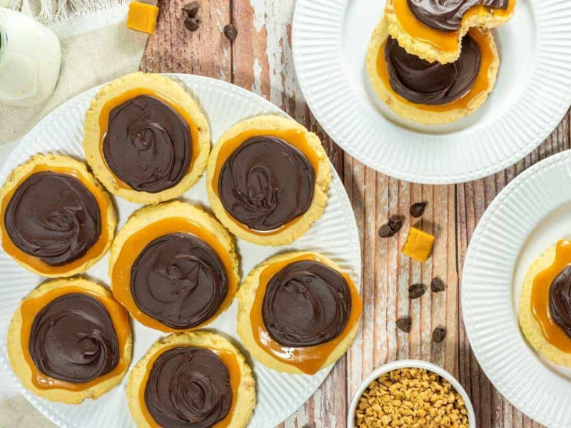 A plate of Copycat Crumbl Twix Cookies, with more cookies on separate plates, caramel pieces, and small chocolate chips nearby on a wooden table.