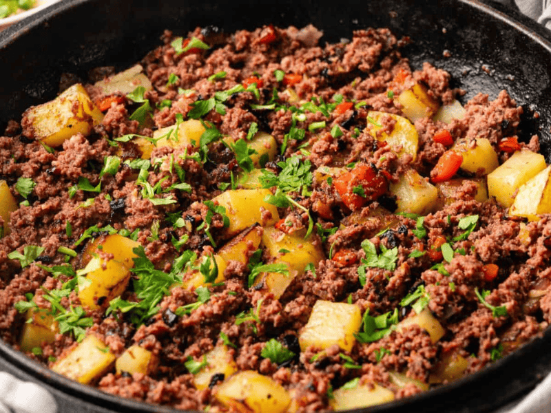 A cast iron skillet filled with cooked ground beef, diced potatoes, red bell peppers, and garnished with chopped parsley.