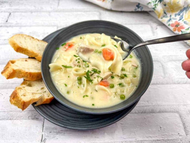 A hand holding a spoonful of creamy chicken noodle soup with vegetables over a bowl, accompanied by slices of bread on a plate.