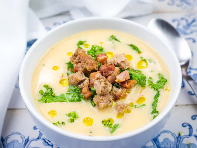 A bowl of Creamy Tuscan Soup with Sausage and Kale set on a blue and white tiled surface with a spoon and napkin beside it.