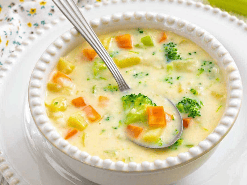 A bowl of creamy vegetable soup with visible broccoli, carrots, and potatoes, served with a spoon on a white plate.