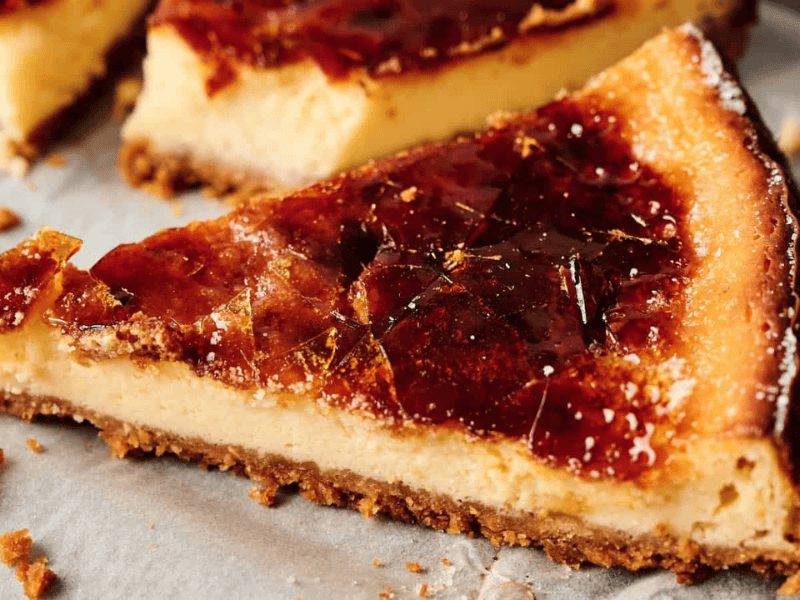 A close-up of a slice of cheesecake with a caramelized sugar topping, resting on parchment paper with a crumbly base visible.