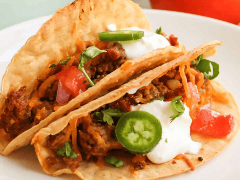 Two tacos with beef, diced tomatoes, jalape&ntilde;o slices, shredded cheese, sour cream, and cilantro on a white plate.