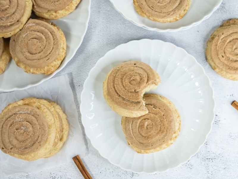 Crumbl Copycat Churro Cookies are arranged on white plates and a napkin. One cookie on a plate has a bite taken out. Two cinnamon sticks are placed nearby on the white surface.