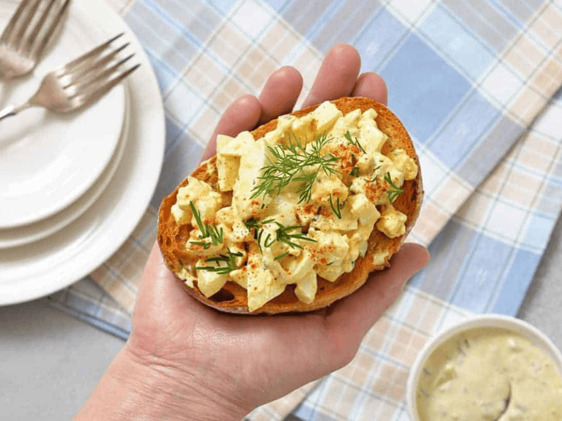 A hand holds a slice of Deviled Egg Toast, garnished with dill and paprika. Three stacked plates with forks and a bowl of sauce are in the background on a checked cloth.