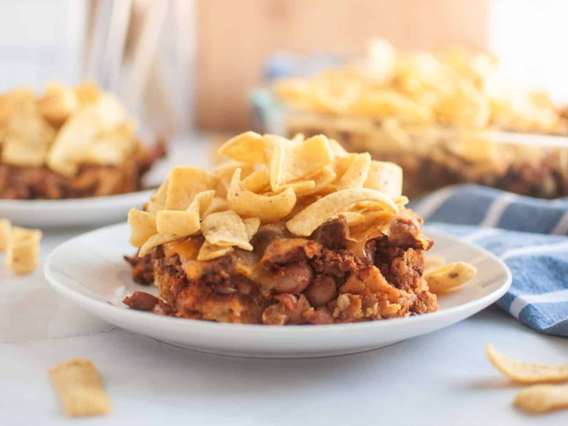 A white plate with a serving of Frito pie, topped with corn chips, is shown on a light surface.