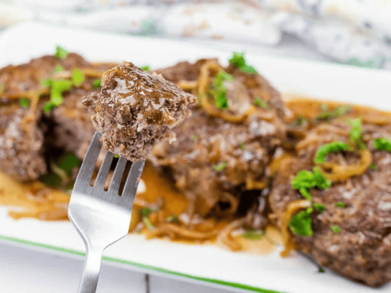 A close-up of a fork holding a piece of cooked Mississippi Salisbury steak with gravy and onions, with more steak pieces and sauce on a white plate in the background.