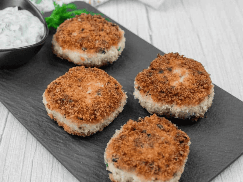 Four Salmon Cakes are arranged on a black slate serving board, accompanied by a small bowl of white sauce with herbs.