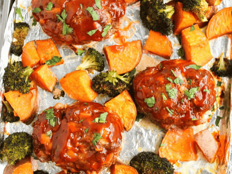 Three mini meatloafs topped with sauce are baked on a foil-lined tray with roasted sweet potato chunks and broccoli florets, garnished with chopped parsley.