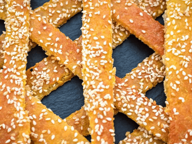 Close-up of several golden sesame breadsticks arranged in a crisscross pattern on a dark surface.