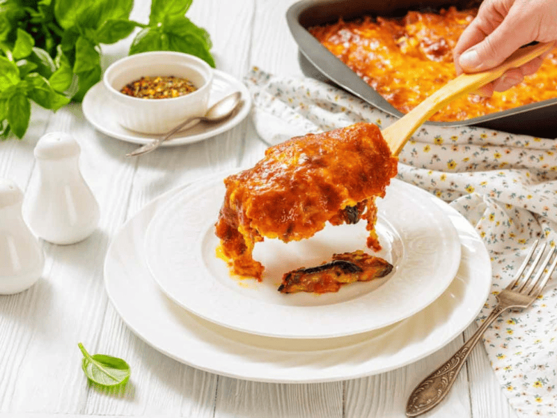 A baked casserole topped with chopped green onions and bacon pieces, with one square being served from the dish using a spatula.