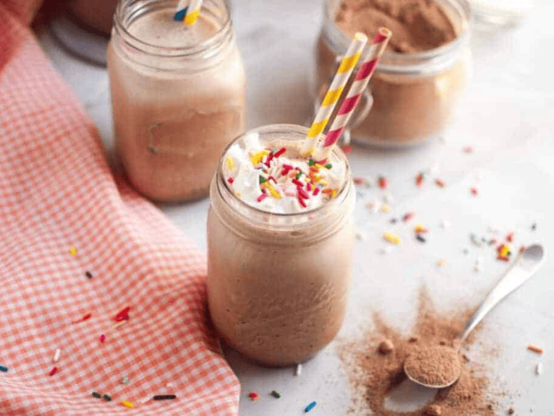 A glass jar filled with a chocolate milkshake topped with whipped cream and colorful sprinkles, with striped straws, surrounded by cocoa powder and a checkered cloth.