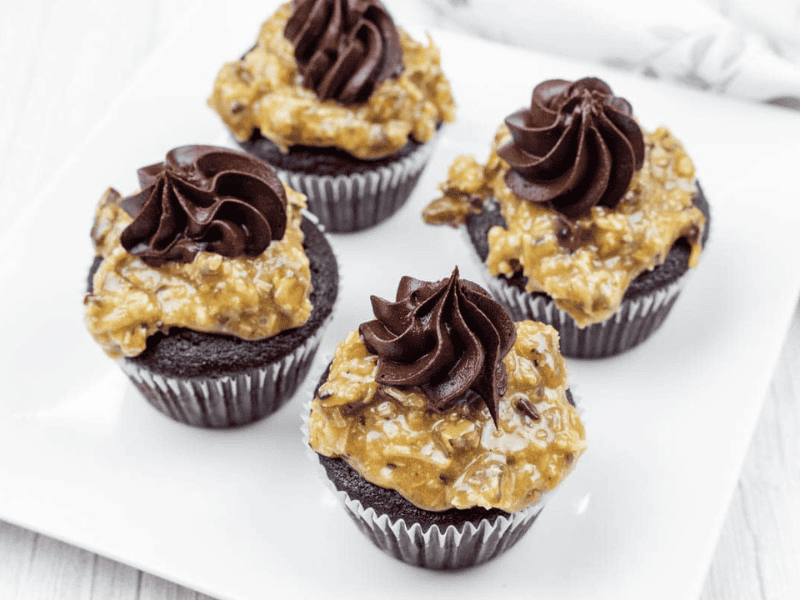 Four German Dark chocolate cupcakes topped with German chocolate coconut-pecan frosting and swirls of chocolate icing, arranged on a white square plate.