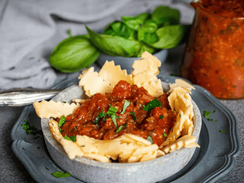 A bowl of tomato-based sauce garnished with chopped herbs, surrounded by broken pieces of flatbread, with fresh basil and a jar of sauce in the background.