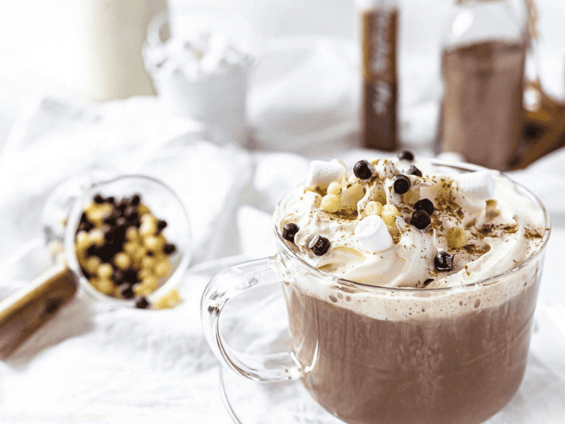 A glass mug of hot chocolate topped with whipped cream, mini marshmallows, and chocolate chips.
