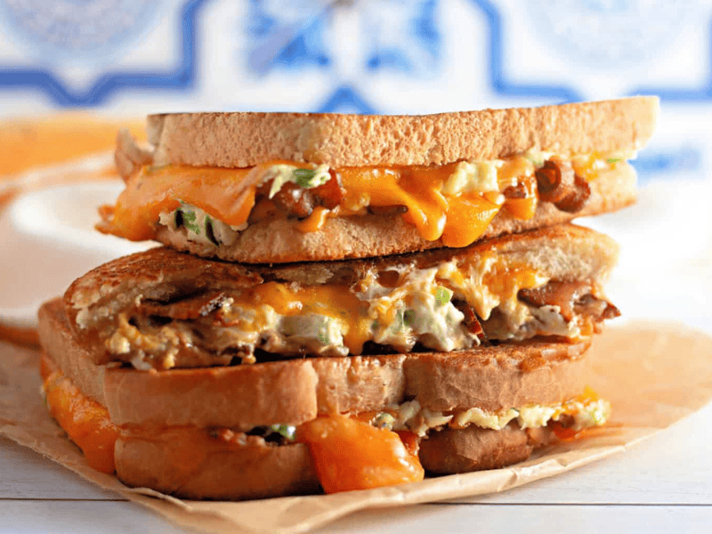 Two Jalape&ntilde;o Popper Grilled Cheese sandwiches with melted cheddar, bacon, and a creamy filling are stacked on parchment paper, with cheese oozing out from the sides.