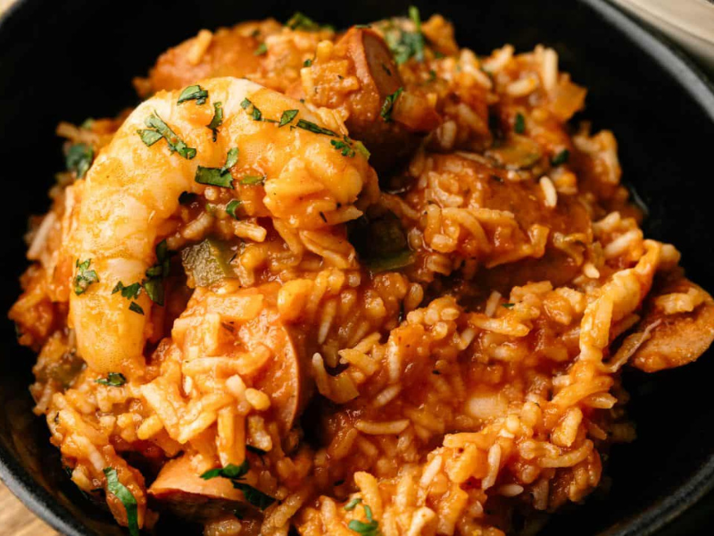 A black bowl filled with jambalaya, featuring shrimp, sausage, rice, and vegetables in a reddish sauce, garnished with chopped herbs.