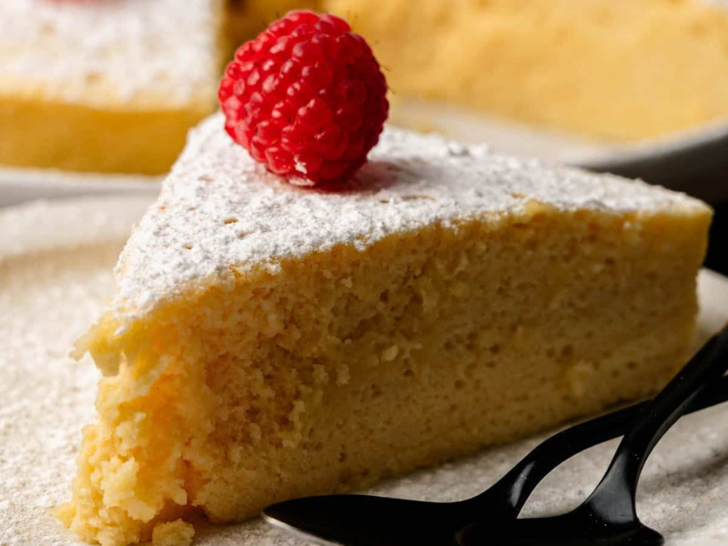 A slice of sponge cake topped with powdered sugar and a raspberry, served on a plate with two black plastic forks.