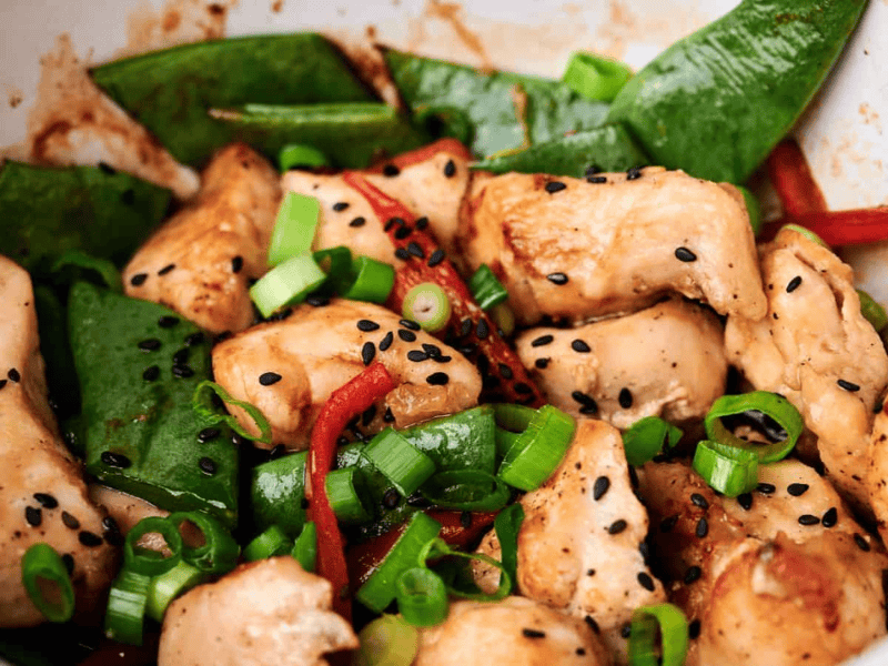 A close-up of a stir-fry dish featuring pieces of chicken, snap peas, red bell peppers, scallions, and black sesame seeds.