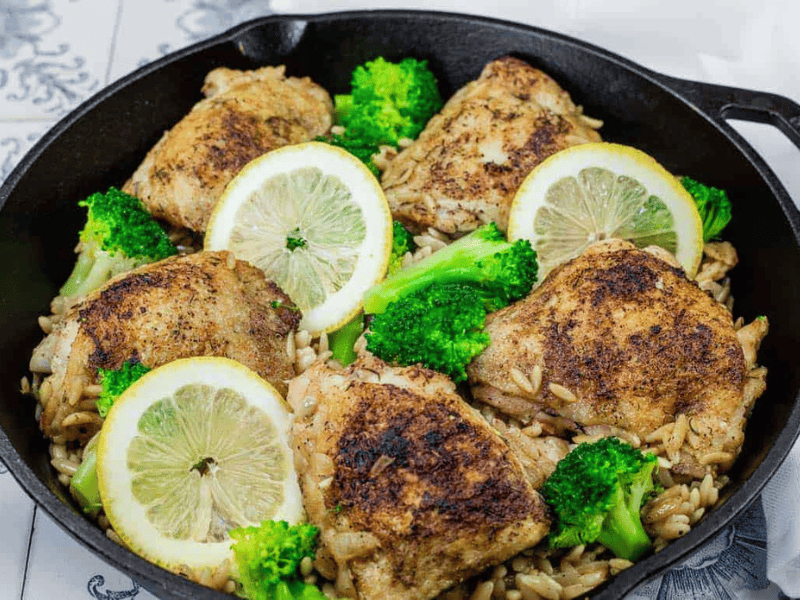 A skillet containing seasoned chicken thighs, broccoli florets, orzo pasta, and lemon slices.