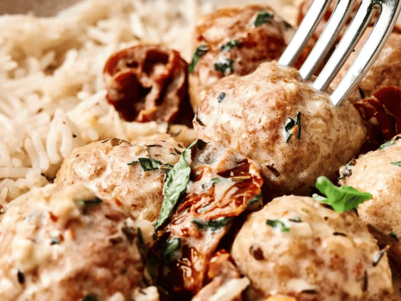 A close-up of meatballs in a creamy sauce with herbs, sun-dried tomatoes, and a side of white rice, with a fork picking up one meatball.