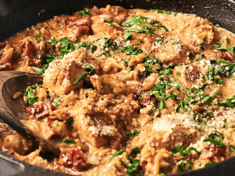 A skillet with creamy chicken pasta featuring spinach, sun-dried tomatoes, and grated cheese, stirred with a wooden spoon.
