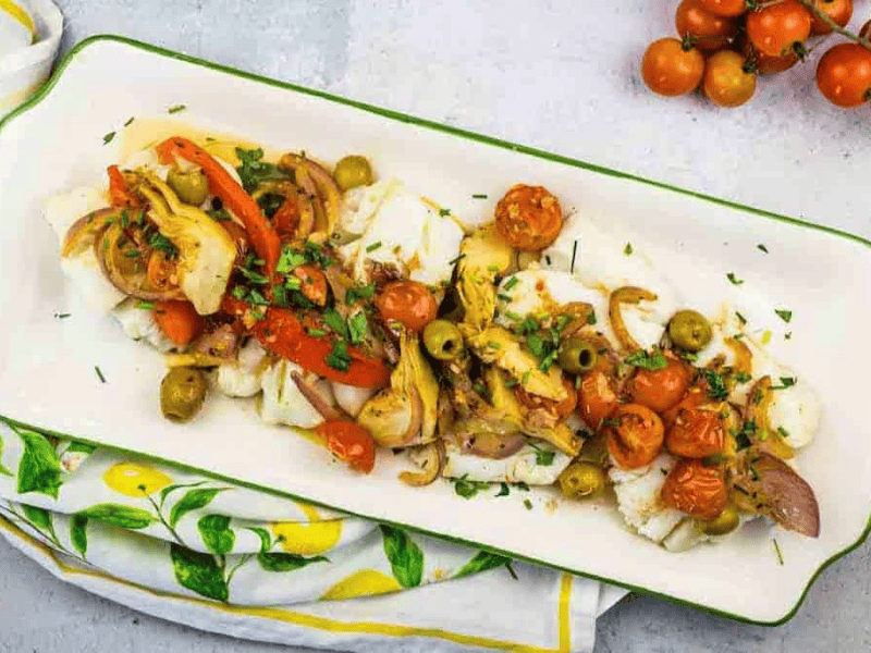 A rectangular plate with Mediterranean Cod placed on a lemon-patterned cloth. A vine of cherry tomatoes is beside the plate.