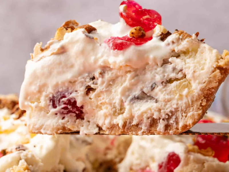 A close-up of a slice of creamy pie with visible cherries, nuts, and a flaky crust, being lifted from the rest of the pie.
