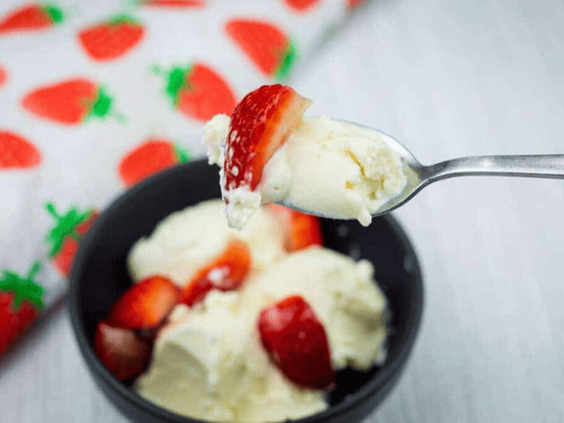 A spoonful of vanilla ice cream and a strawberry held above a bowl with more ice cream and strawberries, next to a strawberry-patterned cloth.