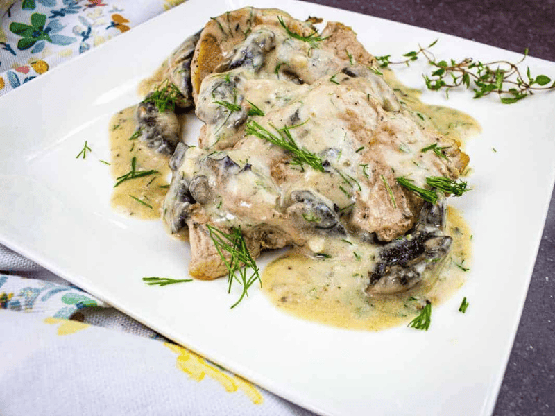 Two pork chops covered in creamy mushroom and herb sauce, garnished with fresh dill, served on a white square plate.