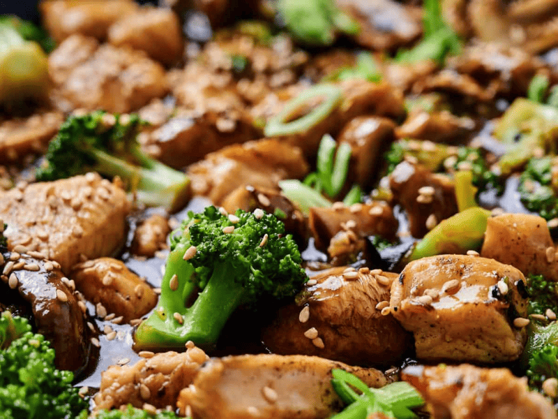 Close-up of a stir-fry dish with chicken, broccoli, mushrooms, and sesame seeds, coated in a soy-based sauce.
