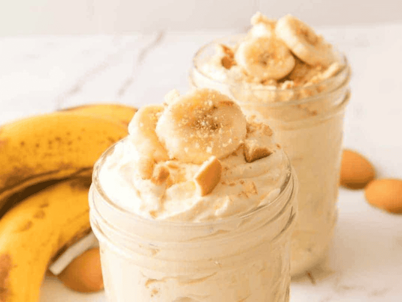 Two jars of banana pudding topped with banana slices and crushed cookies are in the foreground, with bananas and vanilla wafers in the background on a white surface.