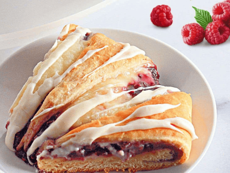 A slice of raspberry crescent ring with white icing on a white plate, with fresh raspberries and a green leaf in the background.