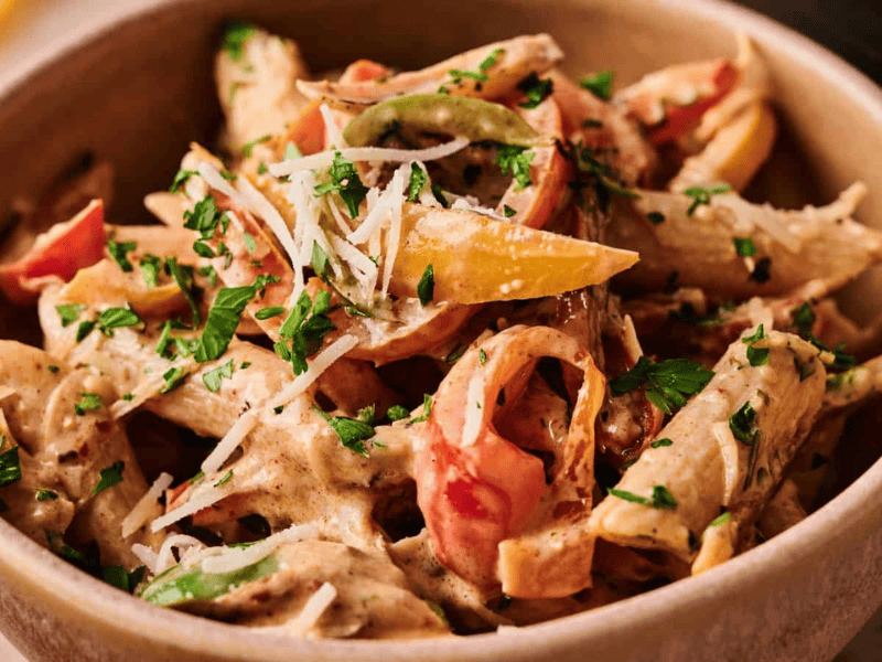 A bowl of penne pasta with creamy sauce, sliced bell peppers, and fresh parsley, topped with grated cheese.