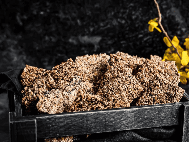 A black tray filled with pieces of sesame seed brittle sits against a dark background with a small yellow flower on the side.