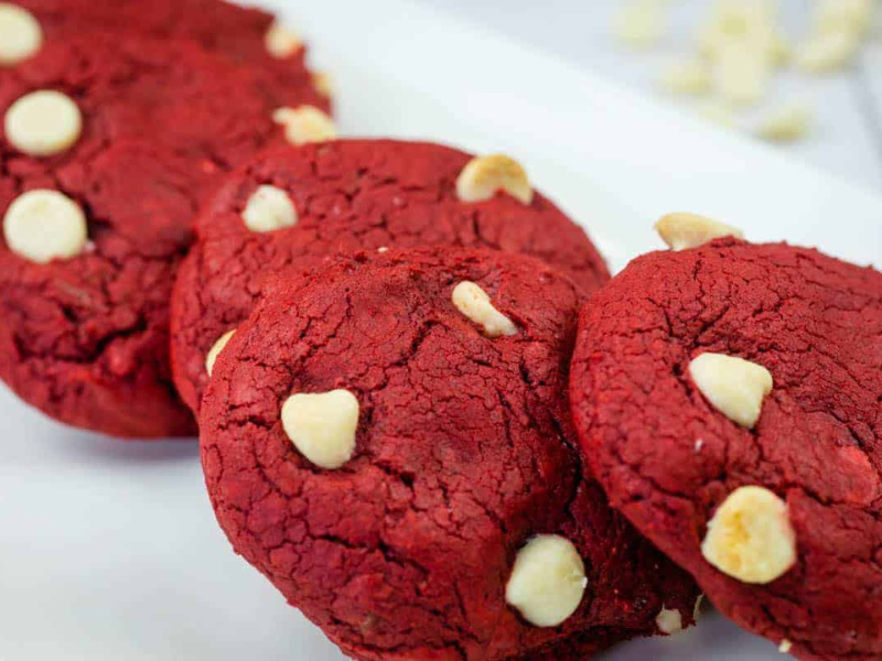 Four red velvet cookies with white chocolate chips on a white rectangular plate.
