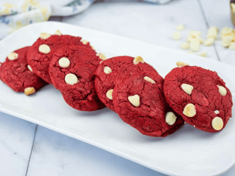 Five red cookies with white chocolate chips are arranged in a row on a rectangular white plate, set on a marble countertop.