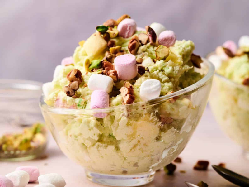 A glass bowl filled with pistachio-flavored dessert topped with mini marshmallows and chopped nuts, with another similar bowl in the background.