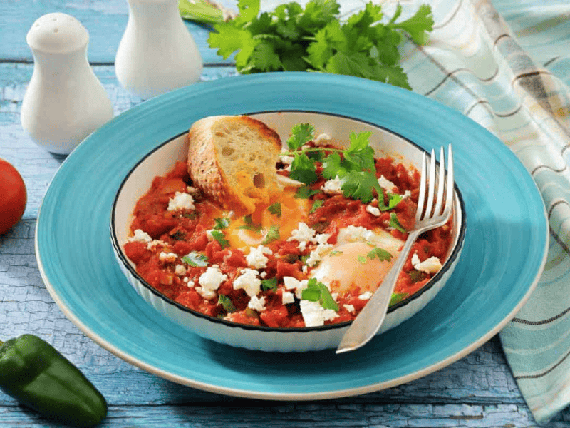 A bowl of shakshuka with poached eggs in tomato sauce, topped with feta and cilantro, served with a slice of toasted bread and a fork on a blue plate.