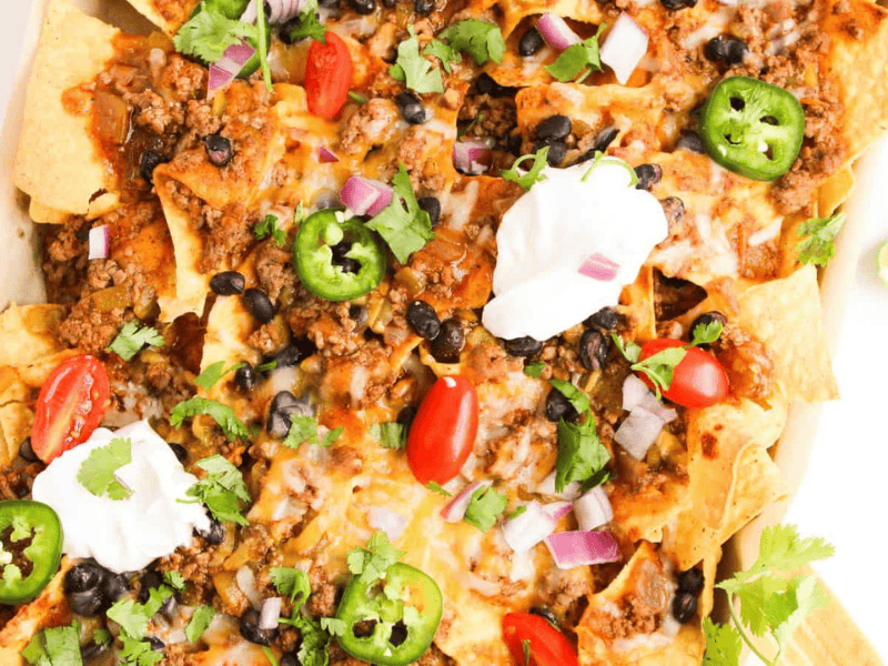 A platter of nachos topped with ground meat, melted cheese, sliced jalape&ntilde;os, cherry tomatoes, black beans, red onions, cilantro, and dollops of sour cream. Lime wedges are on the side.