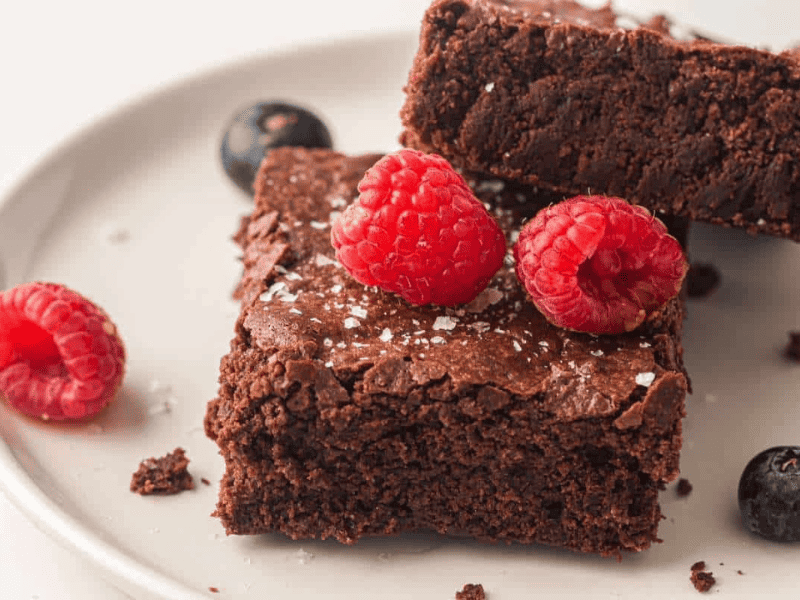 Two Sheet pan chocolate brownies on a white plate, topped with raspberries and a sprinkle of sea salt, with additional raspberries and blueberries on the side.