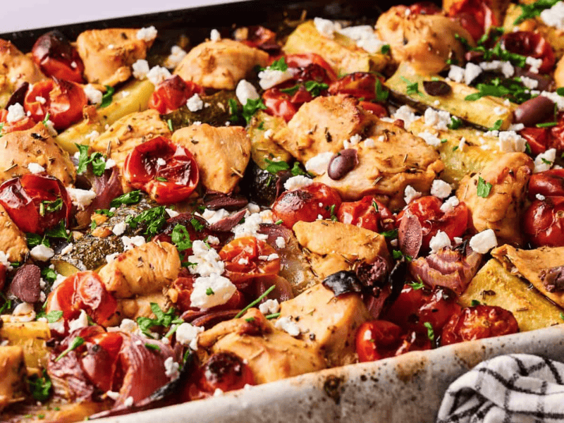 Sheet pan filled with baked chicken pieces, cherry tomatoes, zucchini, olives, and crumbled feta cheese, garnished with herbs.