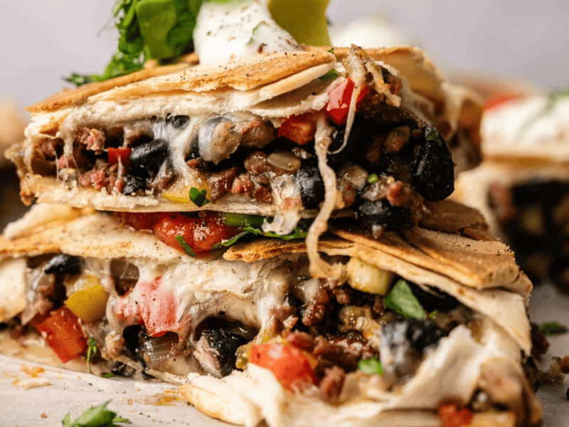 A close-up of stacked quesadilla wedges filled with black beans, ground meat, melted cheese, and diced vegetables, topped with sour cream, guacamole, and chopped herbs.