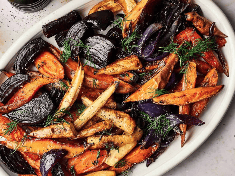 A white dish with roasted root vegetables, including carrots, parsnips, red onions, and sweet potatoes, garnished with fresh dill and thyme.