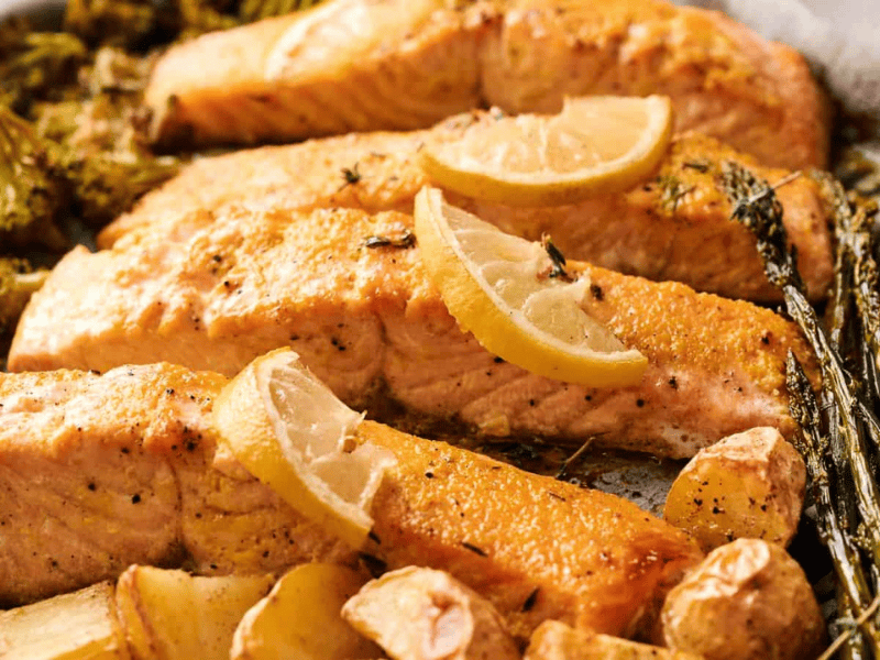 Baked salmon fillets garnished with lemon slices, served with roasted broccoli, asparagus, potatoes, and garlic cloves on a sheet pan.
