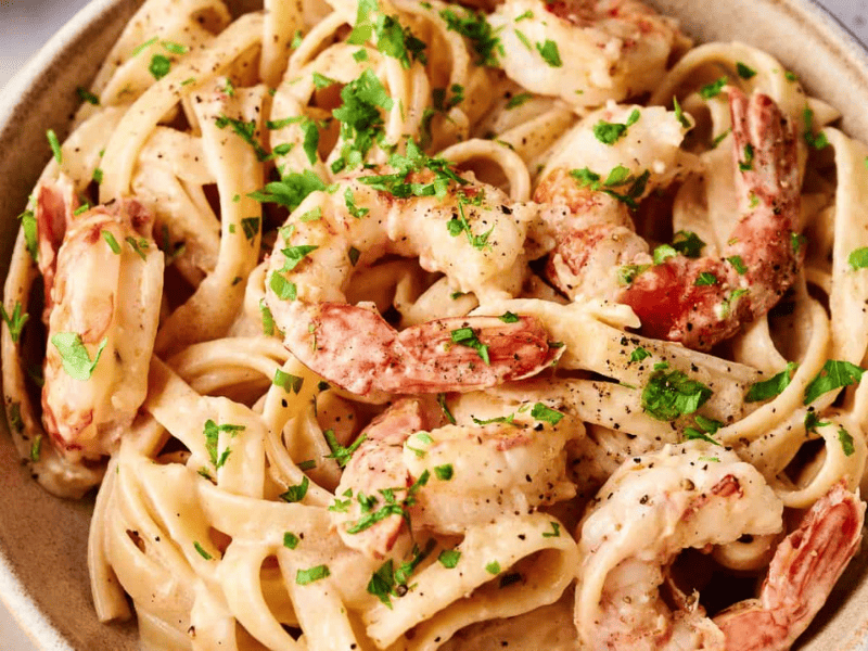 A bowl of fettuccine pasta with shrimp in a creamy sauce, garnished with chopped parsley and black pepper.
