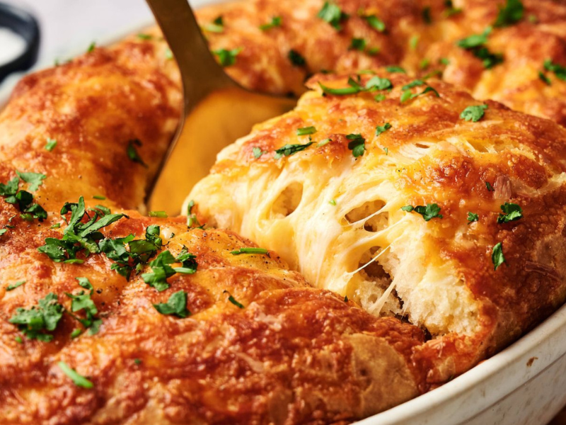 A golden, cheesy bread casserole is being served from a baking dish, with melted cheese stretching and chopped herbs sprinkled on top.