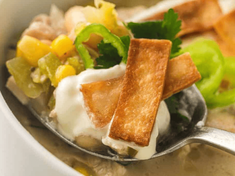 A spoonful of white chili with sour cream, corn, green chili slices, cilantro, and crispy tortilla strips.