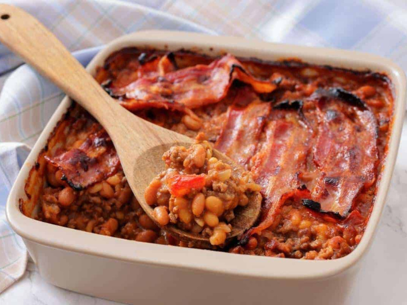A square baking dish filled with Southern Ground Beef & Baked Bean Casserole and topped with strips of cooked bacon; a wooden spoon holds a serving.