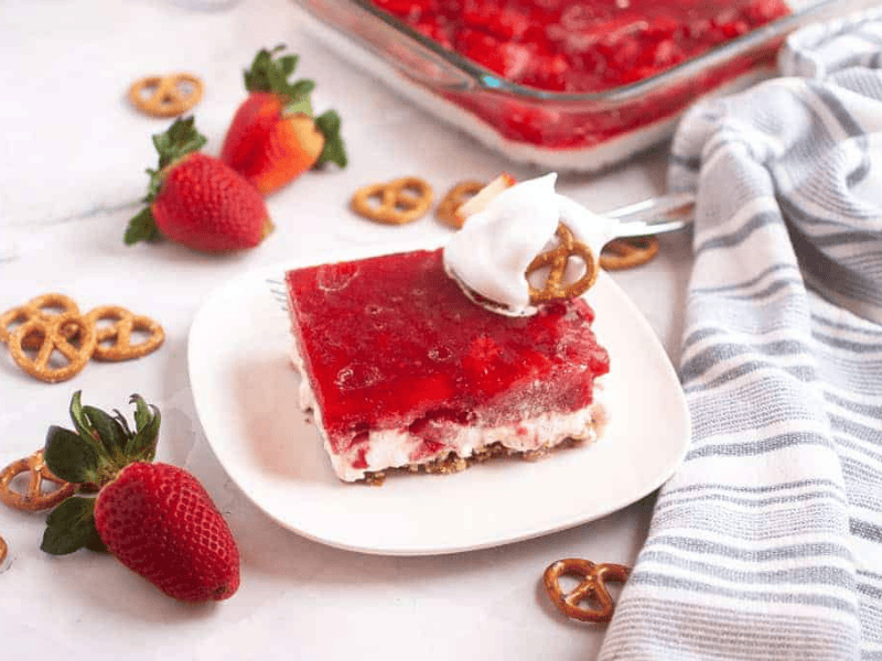 A slice of Strawberry Pretzel Salad topped with whipped cream and a pretzel sits on a white plate.